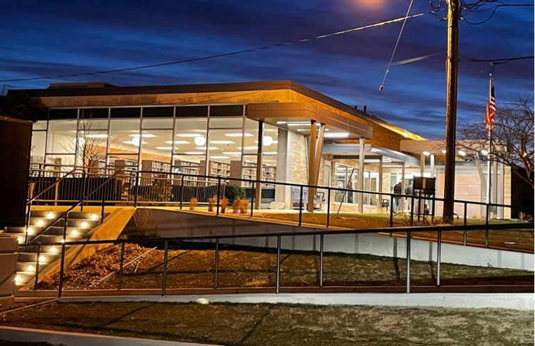 A modern library building with large glass windows is lit up at dusk. Inside, bookshelves and round ceiling lights are visible. An American flag stands outside, and the exterior pathway is illuminated by step lights.